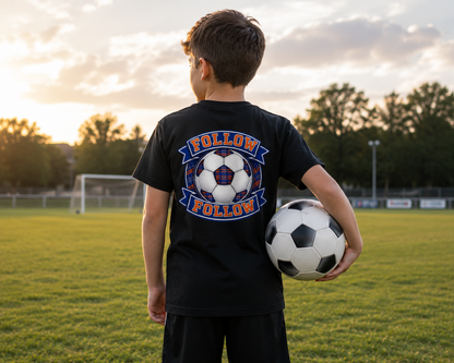 Young football fan wearing a black FOLLOW FOLLOW youth t-shirt with a large back print while holding a football on a grass pitch.