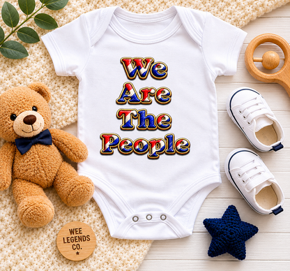 White We Are The People baby bodysuit displayed in a flat lay arrangement with teddy bear, baby shoes, wooden teether and soft nursery accessories on a neutral background.