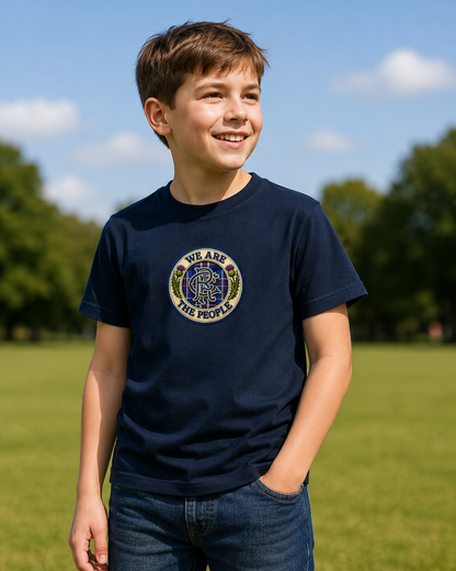Young football supporter wearing a navy We Are The People crest youth t-shirt outdoors on a grassy field, featuring a tartan badge design with Scottish thistles.