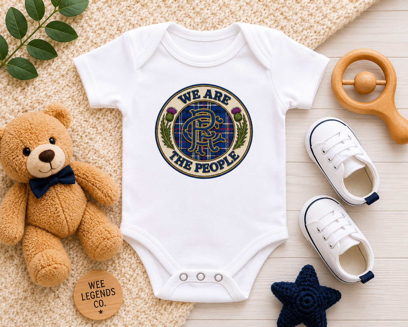 White We Are The People badge baby bodysuit displayed in a flat lay arrangement with teddy bear, baby shoes, wooden rattle and nursery accessories.