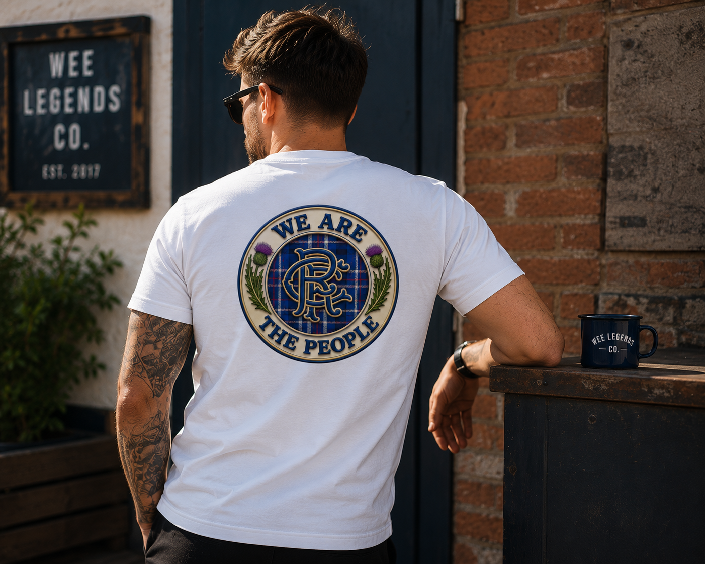 Man relaxing outside a café wearing a white We Are The People Crest back print t-shirt, featuring a large Scottish-inspired tartan crest graphic on the back.