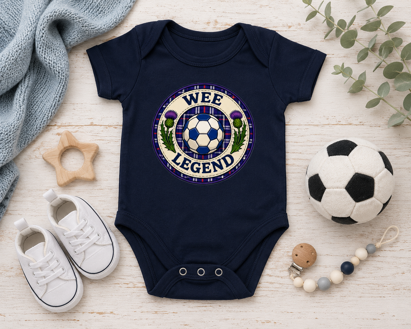 Flat black Wee Legend baby bodysuit styled with baby shoes, football toy and nursery accessories.