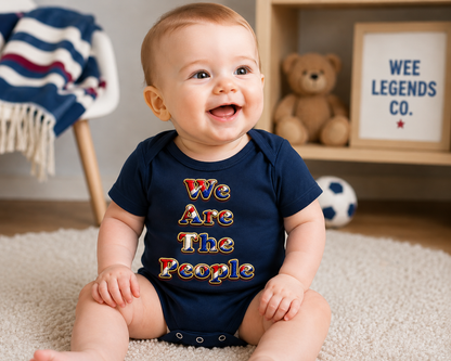 Happy baby wearing a navy We Are The People baby bodysuit featuring a bold red, white and blue slogan design, sitting on a soft rug in a cosy nursery with a teddy bear and football-themed décor.
