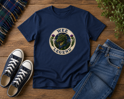 Navy Wee Legend Dragon youth t-shirt displayed on a rustic wooden surface with jeans, trainers, tartan fabric and outdoor adventure accessories.