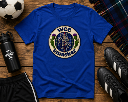 Royal blue Wee Smasher RFC Badge youth t-shirt displayed on a rustic wooden surface with football boots, football, tartan scarf and sports accessories.
