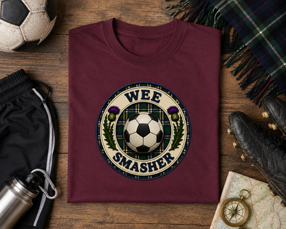 Folded maroon Wee Smasher youth t-shirt displayed on a rustic wooden surface with football boots, football, tartan scarf and outdoor adventure accessories.
