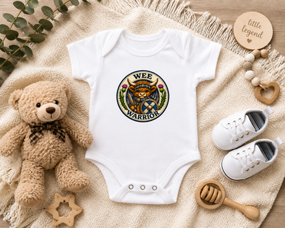 White Wee Warrior baby bodysuit featuring a Highland Cow warrior design displayed in a flat lay arrangement with teddy bear, baby shoes, wooden teether and nursery accessories.