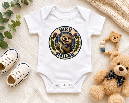White Wee Smiler baby bodysuit with Scottish otter design displayed in a flat lay arrangement with baby shoes, teddy bear, wooden teether and soft neutral nursery accessories.